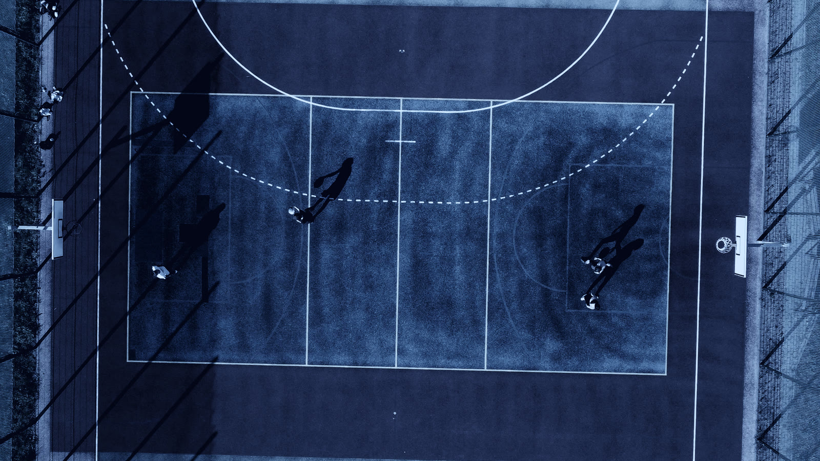 Overhead view of an outdoor court mid game, players casting long shadows across the lines.